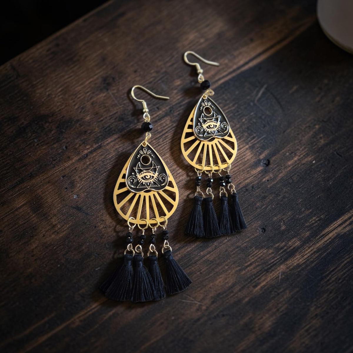 Fury Jewelry & Design mystical eye teardrop dangle earrings in gold-tone metal with black tassels on dark wood.