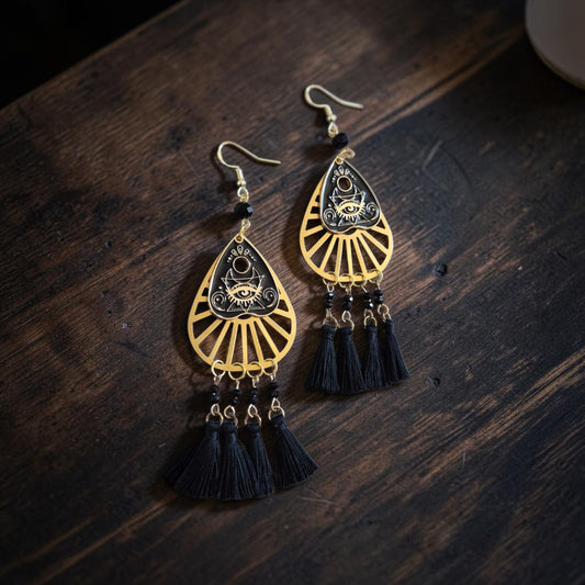 Fury Jewelry & Design mystical eye teardrop dangle earrings in gold-tone metal with black tassels on dark wood.